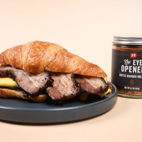 croissant breakfast sandwich with pork belly and scrambled eggs sitting on a dark grey circle plate. To the right of the plate, is a jar of the PS Seasoning The Eye Opener BBQ Rub.