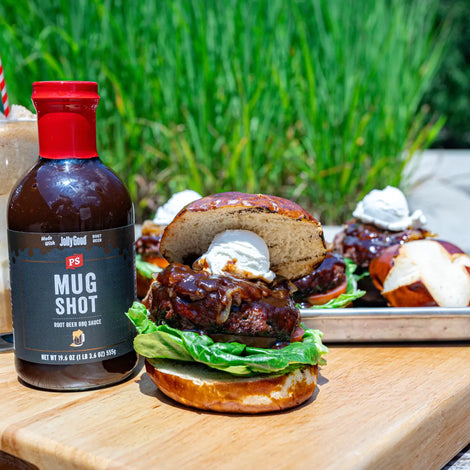 Root Beer Float Burger made with Mug Shot Root Beer BBQ Sauce on a pretzel bun, topped with caramelized onions and scooped goat cheese, served alongside a root beer float and a bottle of PS Mug Shot BBQ Sauce on a wooden board outdoors.