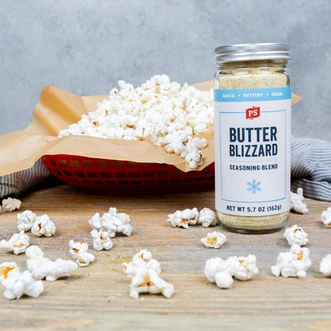 Basket of freshly popped popcorn seasoned with PS Seasoning Butter Blizzard, with popcorn scattered on a wooden surface and a jar of Butter Blizzard seasoning in the foreground. Simple garlic butter snack made with stovetop popcorn.