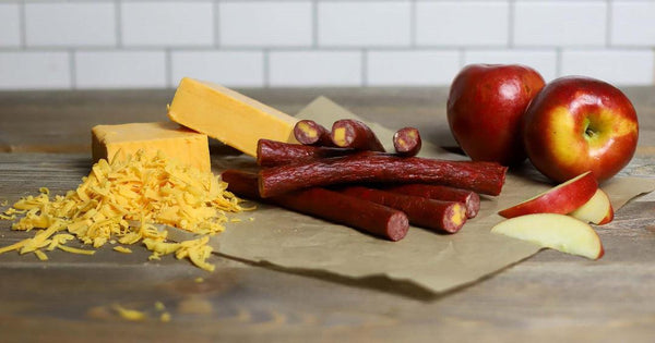 Cheese and apple beef sticks made with high temp cheddar cheese. Beef sticks are laying next to apples and a block of shredded cheese.