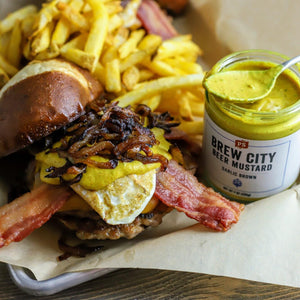 A burger next to an open jar of Brew City Beer Mustard, which has a spoon in it