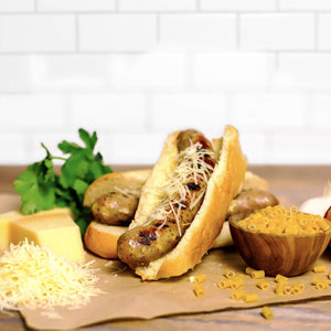 Two Alfredo Brat's with cheese on top next to a small bowl of noodles and shredded cheese.