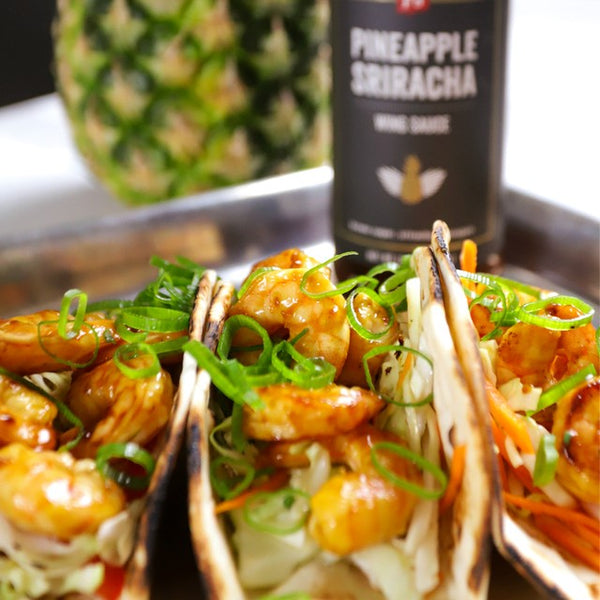 3 Pineapple Sriracha Shrimp Tacos in front of a bottle of Pineapple Sriracha and a pineapple.