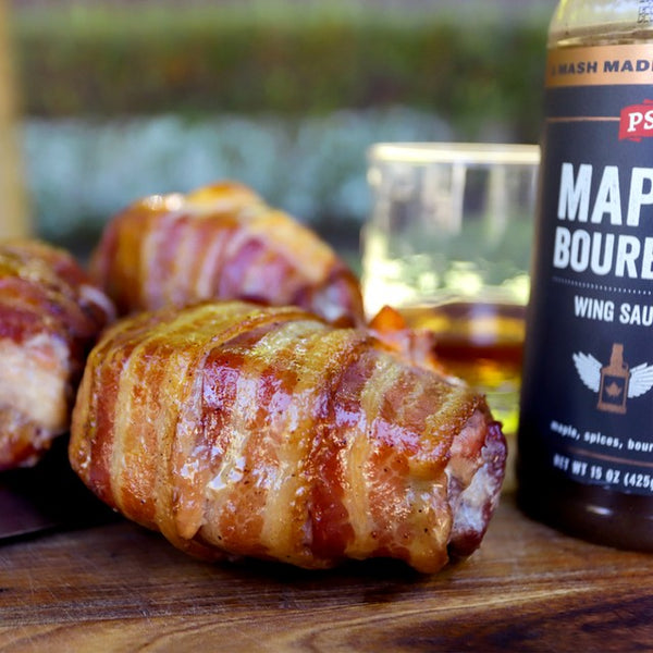 Apple stuffed pork chops covered in bacon next to a bottle of Maple Bourbon Wing Sauce.