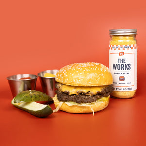 The Works Burger Seasoning is next to a double smash burger, two pickles, a cup of ketchup, and mustard.