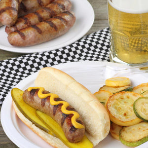 Beer Brat on a bun with pickles and mustard. Next to it is veggies, with a beer and a plate of brats behind it.