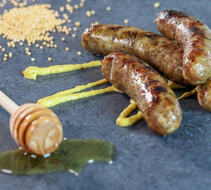 Honey Mustard Brats piled on one another with a mustard drizzle on them. To the left is a honey stick with honey dripping on the table, and Honey Mustard seasoning in the back left hand corner.