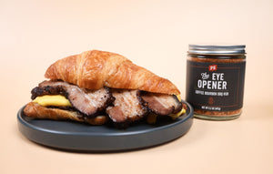 croissant breakfast sandwich with pork belly and scrambled eggs sitting on a dark grey circle plate. To the right of the plate, is a jar of the PS Seasoning The Eye Opener BBQ Rub.