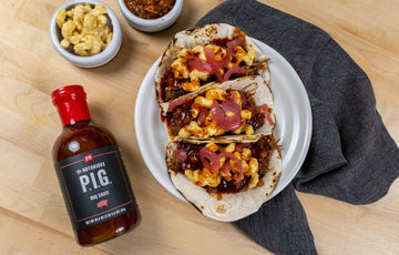 three pulled pork tacos with mac and cheese and pickled onions on top sitting on a white, circular plate. There is a bottle of Notorious PIG BBQ sauce to the left of the plate.
