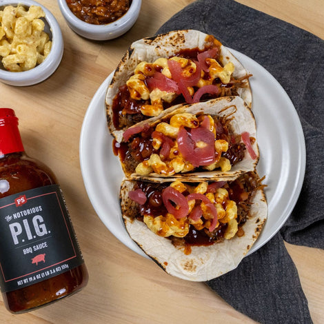 three pulled pork tacos with mac and cheese and pickled onions on top sitting on a white, circular plate. There is a bottle of Notorious PIG BBQ sauce to the left of the plate.