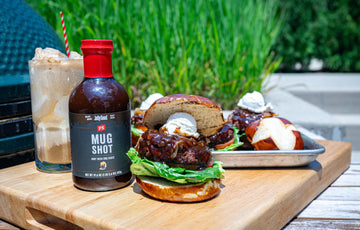 Root Beer Float Burger made with Mug Shot Root Beer BBQ Sauce on a pretzel bun, topped with caramelized onions and scooped goat cheese, served alongside a root beer float and a bottle of PS Mug Shot BBQ Sauce on a wooden board outdoors.