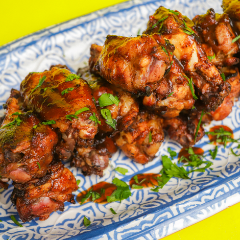 A platter of grilled chamoy chicken wings garnished with fresh chopped cilantro and drizzled with chamoy sauce, served with lime wedges on a decorative blue-and-white plate against a bright yellow background.