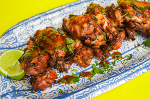 A platter of grilled chamoy chicken wings garnished with fresh chopped cilantro and drizzled with chamoy sauce, served with lime wedges on a decorative blue-and-white plate against a bright yellow background.