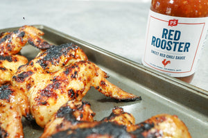 Close-up of grilled chicken wings glazed with Red Rooster Sweet Red Chili Sauce on a baking tray, with a jar of the sauce visible in the background.