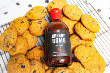 Bottle of PS Seasoning Cherry Bomb BBQ Sauce resting on a wire rack surrounded by freshly baked cherry chocolate chip cookies. Sweet and smoky cookies infused with barbecue sauce, dried cherries, and chocolate chips.