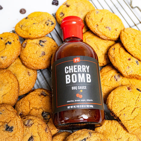 Bottle of PS Seasoning Cherry Bomb BBQ Sauce resting on a wire rack surrounded by freshly baked cherry chocolate chip cookies. Sweet and smoky cookies infused with barbecue sauce, dried cherries, and chocolate chips.