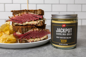 Smoked corned beef Reuben sandwich stacked on rye bread with melted Swiss cheese and sauerkraut, served with kettle chips beside a jar of PS Seasoning Jackpot Carolina Gold Honey Mustard BBQ Rub.