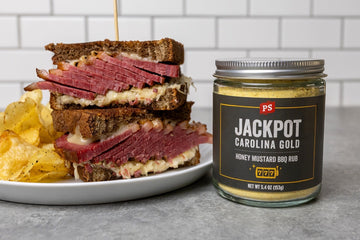 Smoked corned beef Reuben sandwich stacked on rye bread with melted Swiss cheese and sauerkraut, served with kettle chips beside a jar of PS Seasoning Jackpot Carolina Gold Honey Mustard BBQ Rub.