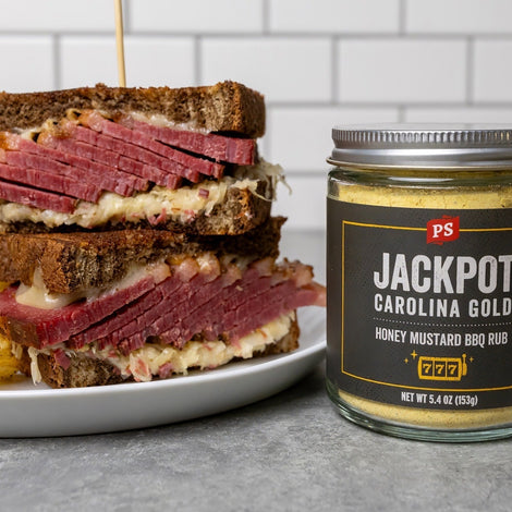 Smoked corned beef Reuben sandwich stacked on rye bread with melted Swiss cheese and sauerkraut, served with kettle chips beside a jar of PS Seasoning Jackpot Carolina Gold Honey Mustard BBQ Rub.