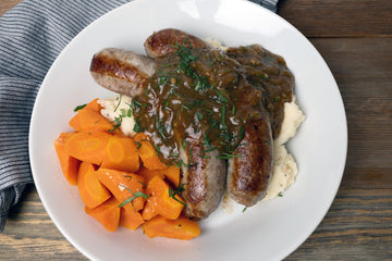 Onion and garlic bratwurst served over creamy mashed potatoes with Guinness onion gravy, garnished with herbs and paired with sliced carrots. Hearty sausage dinner featuring PS Seasoning Chop Shop seasoning.