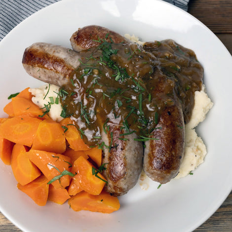 Onion and garlic bratwurst served over creamy mashed potatoes with Guinness onion gravy, garnished with herbs and paired with sliced carrots. Hearty sausage dinner featuring PS Seasoning Chop Shop seasoning.