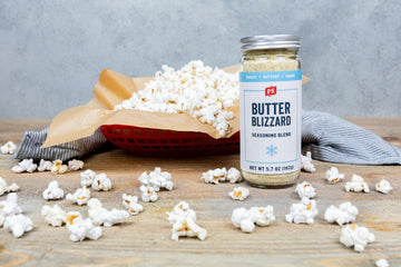 Basket of freshly popped popcorn seasoned with PS Seasoning Butter Blizzard, with popcorn scattered on a wooden surface and a jar of Butter Blizzard seasoning in the foreground. Simple garlic butter snack made with stovetop popcorn.