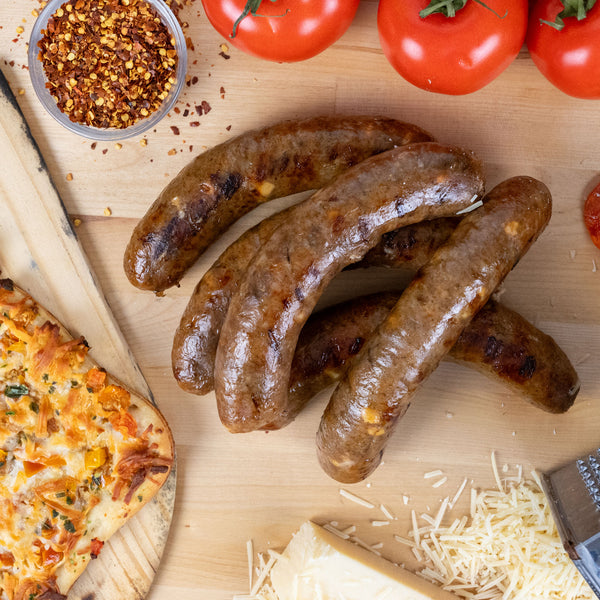 Wood-Fired Pizza Brat next to a flatbread pizza, graded cheese, tomatoes, and pepper flakes.