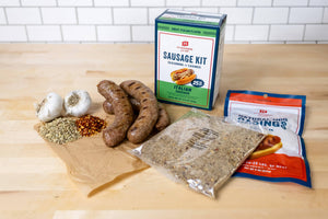 A sausage kit ingredients laid out on a light wood countertop, including a box of sausage links, a bag of seasoning mix, a package of breading sauce, garlic bulbs, and red pepper flakes, with a white subway tile backsplash behind.