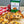A slice of sausage pizza being lifted with a metal spatula, with a sliced pizza, a red-and-white checkered cloth, and a green and white 'Sausage Kit' box in the background on a wooden surface.