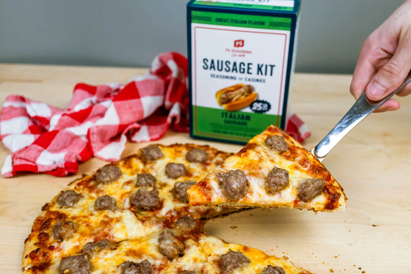 A slice of sausage pizza being lifted with a metal spatula, with a sliced pizza, a red-and-white checkered cloth, and a green and white 'Sausage Kit' box in the background on a wooden surface.