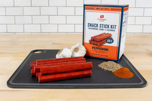 A Pepperoni Snack Stick Kit in a black cutting board with garlic, cured pepperoni sticks, and seasoning piles on a wooden surface against a white subway tile background.