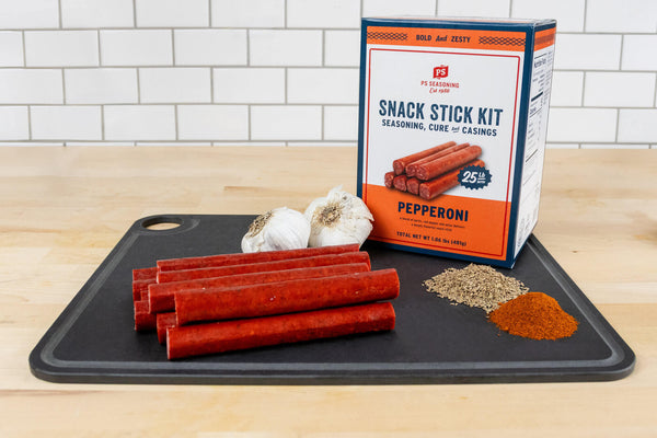 A Pepperoni Snack Stick Kit in a black cutting board with garlic, cured pepperoni sticks, and seasoning piles on a wooden surface against a white subway tile background.