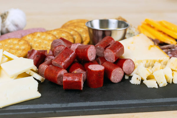 A charcuterie-style food platter on a black serving board, featuring sliced red sausage, crackers, cheese wedges and cubes, and a small metal cup on a light wood surface.