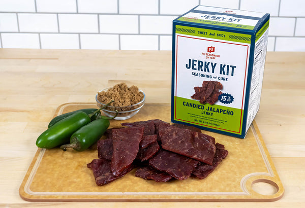 A packaged jerky kit box labeled 'Candied Jalapeño Jerky' stands on a wooden cutting board next to sliced jerky pieces