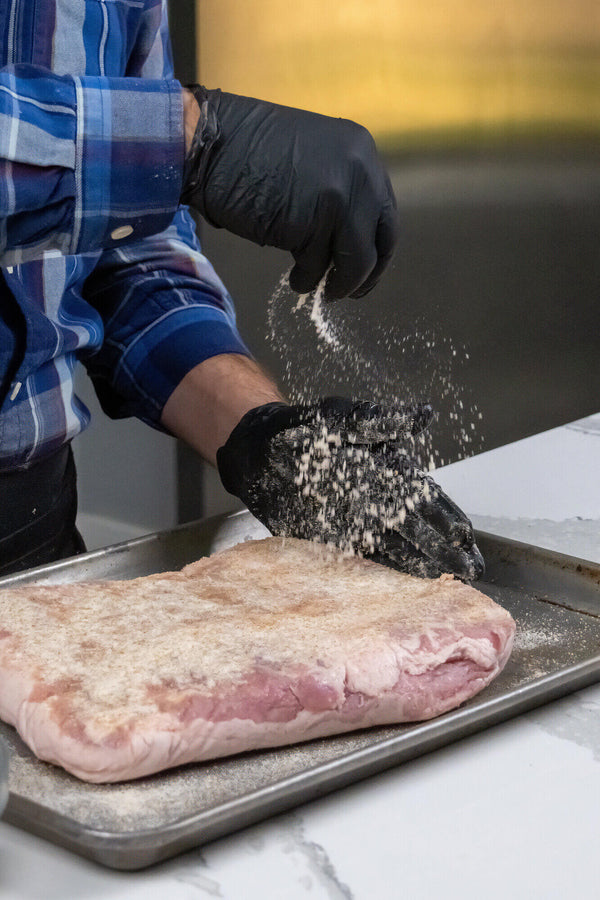 A person wearing black gloves sprinkles coarse white seasoning over a raw cut of pork belly resting on a metal baking sheet.