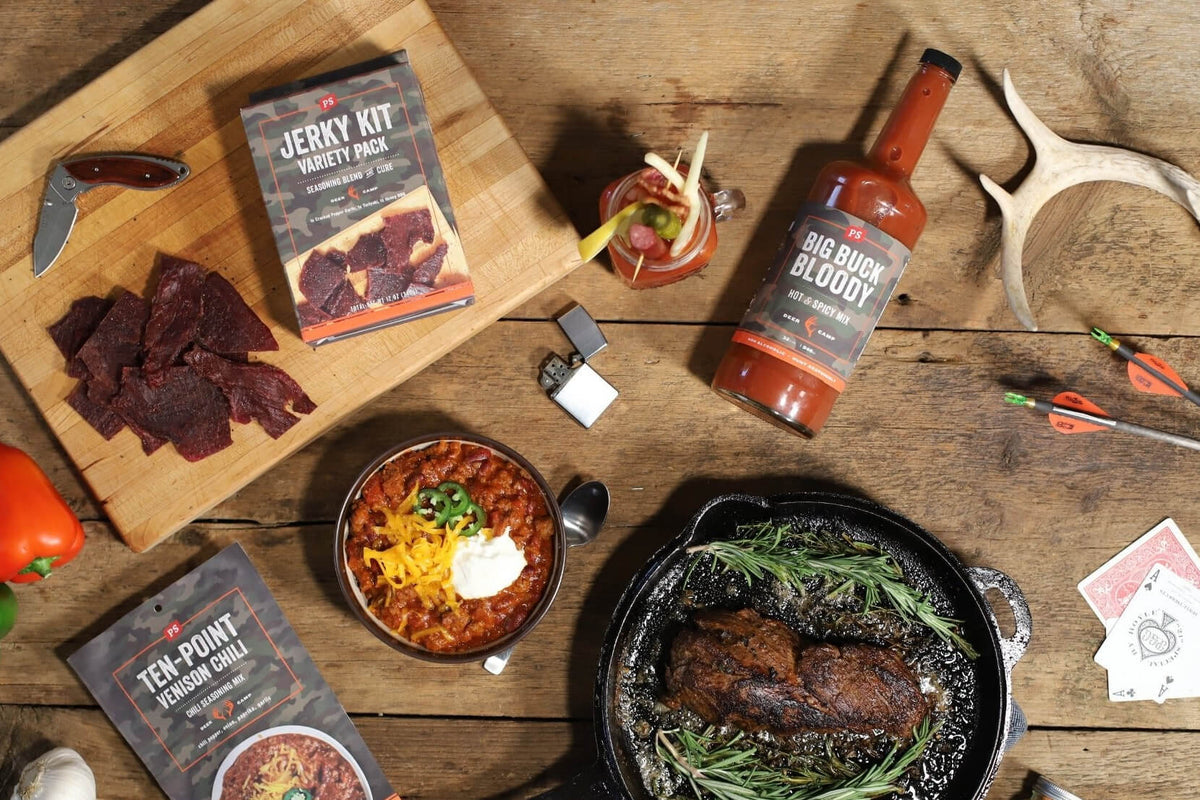 A styled flat-lay of a barbecue food kit on a rustic wooden table, featuring a boxed jerky kit, a bottle of Big Buck Bloody 
Mary Mix, packaged ten-point chili mix