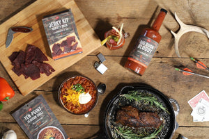 A styled flat-lay of a barbecue food kit on a rustic wooden table, featuring a boxed jerky kit, a bottle of Big Buck Bloody 
Mary Mix, packaged ten-point chili mix