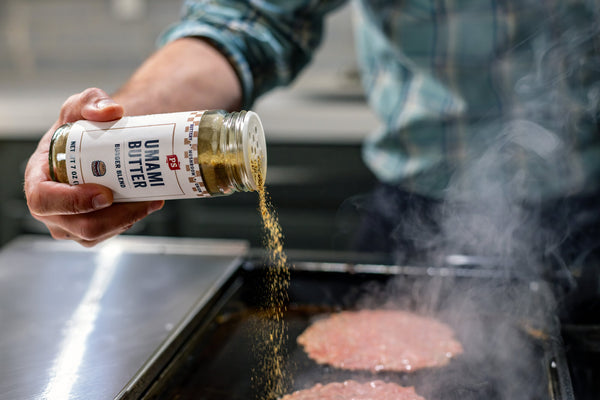 Person seasoning burgers with Umami Bitter seasoning on a grill