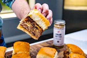 Hand holding a sliced sandwich with a jar of Umami Butter on a table