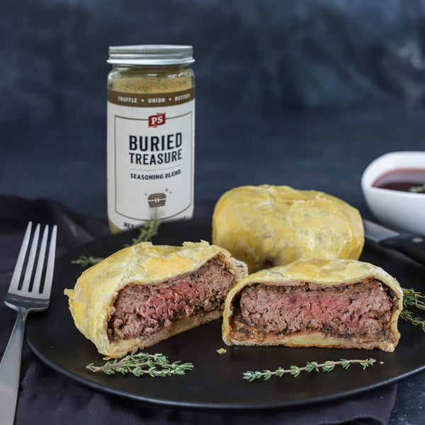 A jar of PS Buried Treasure Seasoning Blend stands behind plated beef pie halves on a black plate, with a fork on the left and a small bowl on the right, against a dark textured background.