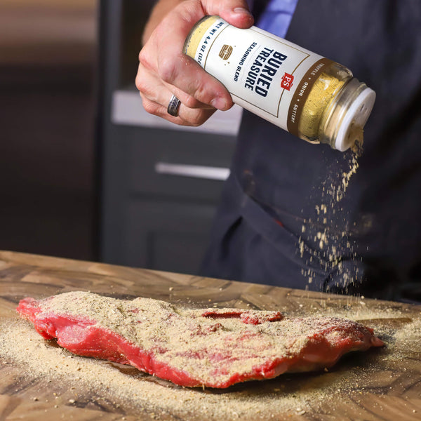A person in a dark apron sprinkles Burled Preside seasoned salt over a raw cut of beef on a wooden cutting board.