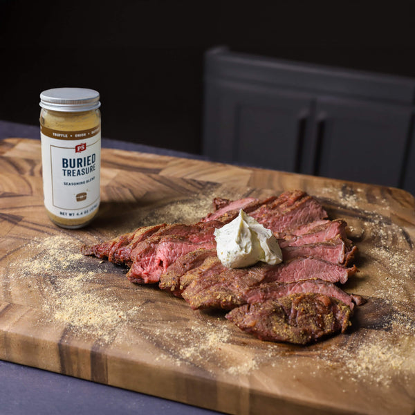 A jar of Burles Treasure Seasoning Blend sits on a wooden cutting board next to sliced cooked meat topped with a dollop of white sauce, with coarse seasoning scattered around.