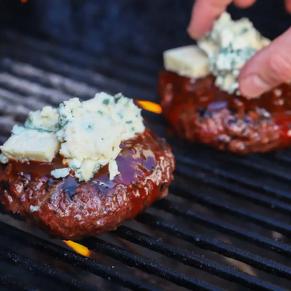 A burger on the grill with Cherry Bomb on it covered in cheese.