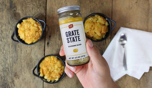 Person holding a jar of 'Grate State' seasoning blend over macaroni and cheese on a wooden table.
