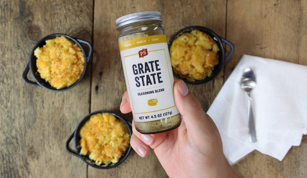 Person holding a jar of 'Grate State' seasoning blend over macaroni and cheese on a wooden table.