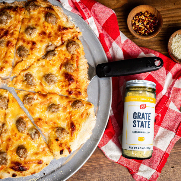 Pizza with sausage pieces on a wooden table with 'Grate State' seasoning blend nearby.