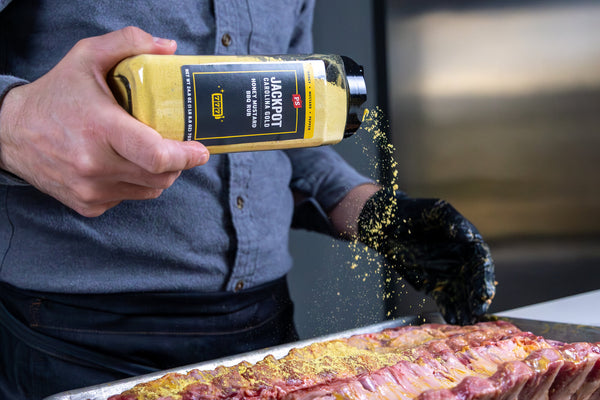 Person seasoning a rib rack with yellow spice mix from a bottle labeled 'Jacques'.