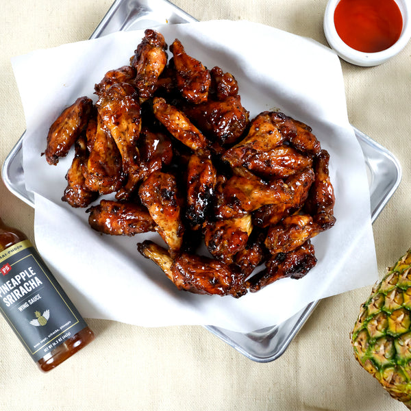 Pineapple sriracha BBQ sauce used on chicken wings, next to a bottle of Pineapple Sriracha and a pineapple. 