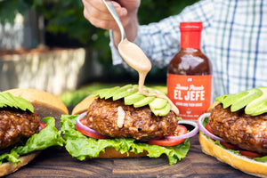 Person adding sauce to a burger with 'El Jefe' bottle in the background