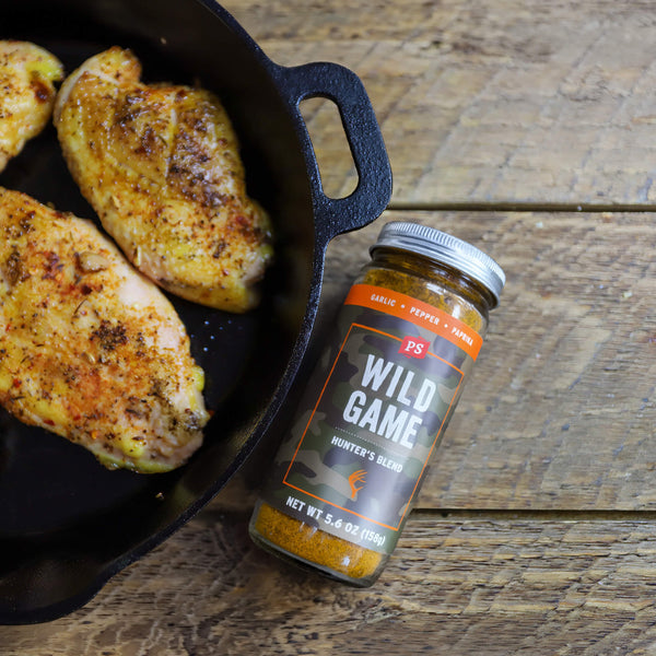 Skillet with seasoned chicken and a jar of P.S. Wild Game seasoning on a wooden surface